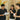 Three people in black shirts with ANGKAN logo, one holding a drink and paper, in a casual setting.